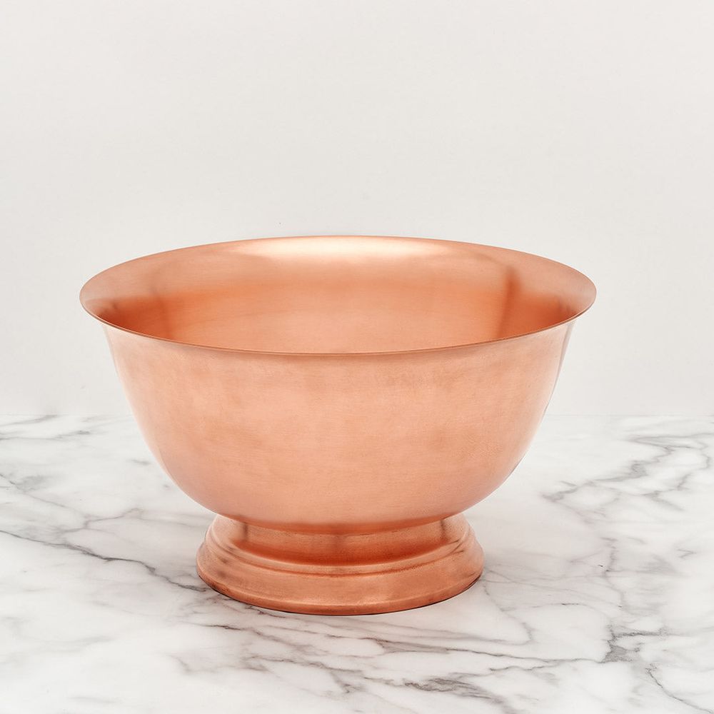Polished copper footed bowl with a reflective rosy-orange hue resting on a white marble surface featuring gray veining